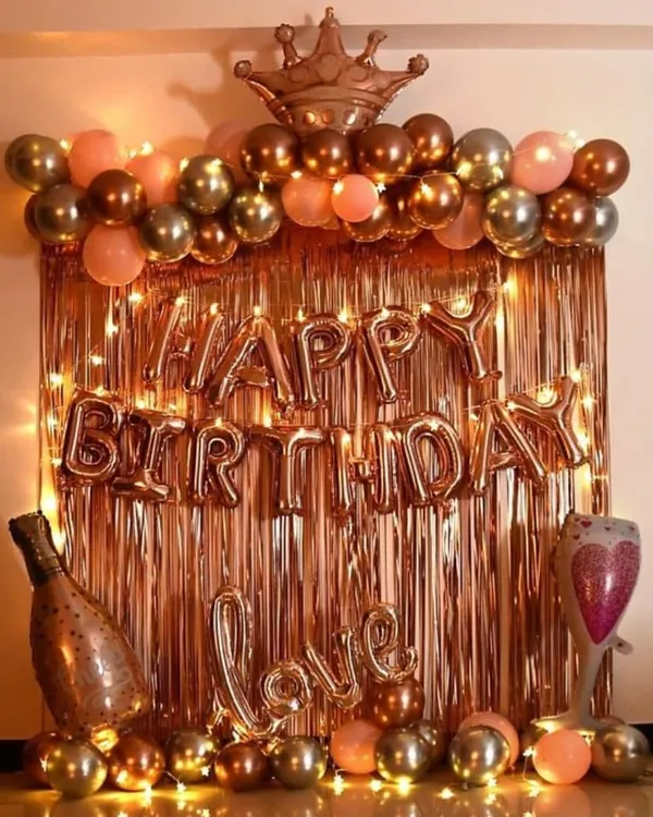 A very popular birthday decoration in a rose gold and gold theme. This setup includes a shiny foil curtain, 'Happy Birthday' letters, a balloon arch topped with a crown, and other fun balloons like a champagne bottle and a 'Love' sign.
