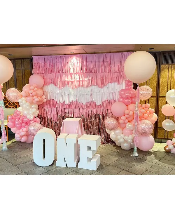 This is the full view of our fringe wall installation for a first birthday. The setup includes large 'ONE' marquee letters, two large balloon columns, and the hand-built pink, white, and rose gold fringe backdrop, creating a beautiful focal point.