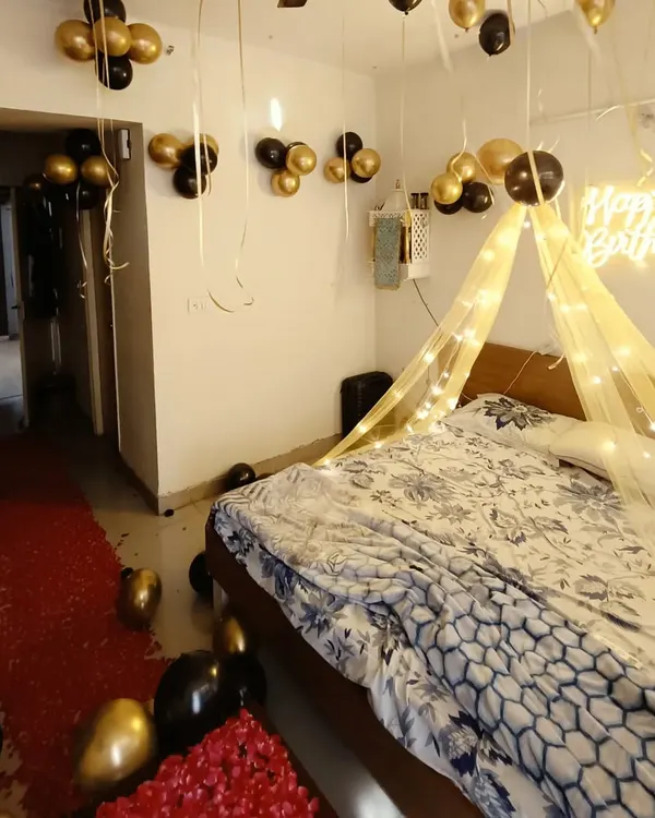 A romantic birthday room surprise with a black and gold theme. The setup includes a bed canopy with fairy lights, a neon sign, a red rose petal pathway, and balloons on the ceiling.