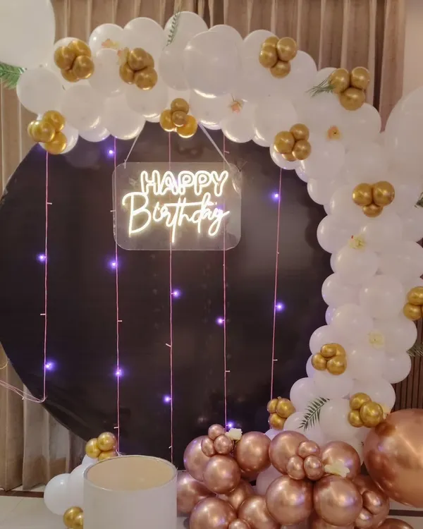 A sophisticated decoration featuring a black circular backdrop. I framed it with a white, rose gold, and gold balloon arch and added a "Happy Birthday" neon sign with hanging fairy lights.