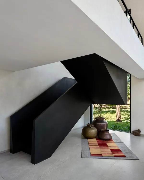 This staircase in the Mango Orchard House is designed as a bold, black, folded-metal sculpture. It creates a powerful visual statement against the light-colored walls, with a large window at the base framing a view of the garden.