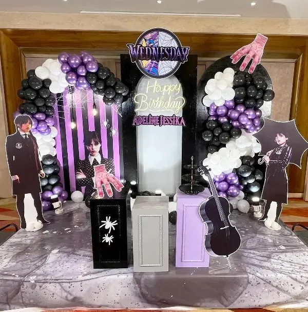 A complete 'Wednesday' themed birthday party setup, featuring character cutouts of Wednesday Addams, a cello prop, and the disembodied hand, Thing. The backdrop includes a custom neon sign, themed logo, and balloon arches in black, white, and purple for a spooky, stylish event in Bangalore.