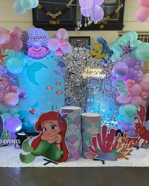 A complete 'Under the Sea' birthday party setup for Viyana. The scene is set with a silver shimmer wall, character cutouts of Ariel and Flounder, and custom props like coral and a treasure shell.