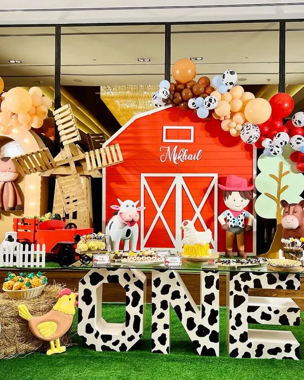 We had so much fun creating this complete barnyard experience for little Mikhail's first birthday. This setup features a classic red barn backdrop, a rustic windmill, life-sized farm animal props like a cow and sheep, and custom cow-print 'ONE' letters for the perfect photo op.