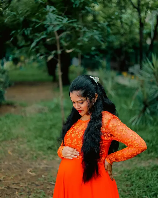 A quiet moment of connection in a peaceful park setting. This outdoor maternity portrait captures the mother-to-be in a vibrant orange dress, surrounded by natural greenery.