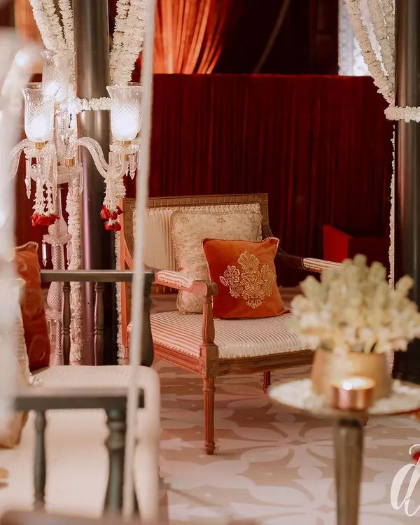 A close-up of a seating area from a royal-themed reception. The decor features elegant candelabras draped with jasmine, set against a backdrop of rich, red velvet curtains.