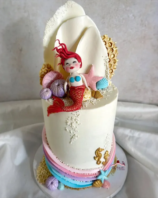 This mermaid cake is a more artistic take on the theme, featuring a hand-sculpted mermaid with vibrant red hair resting against a large, white chocolate seashell. Edible sand, seahorses, and coral details complete the underwater scene.