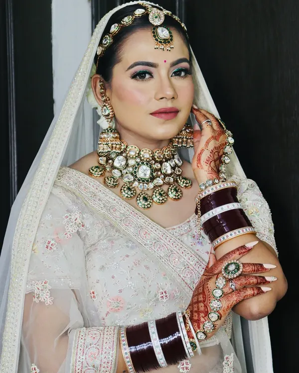 A stunning modern bride in a white lehenga. The makeup features a pop of green eyeliner, soft pink tones, and a flawless base, complemented by heavy Kundan jewellery.