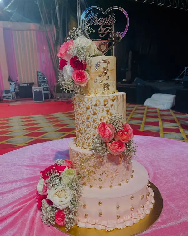 A four-tiered masterpiece fit for a royal celebration. This cake combines different textures, from smooth pink buttercream to intricate gold piping, all brought together with cascades of fresh pink and white roses.