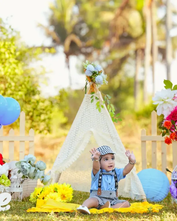 A baby boy enjoys an outdoor photoshoot in a beautiful, sunny field. The setup includes a lace teepee, colorful flowers, and balloons, creating a perfect picnic-like atmosphere for his first birthday.