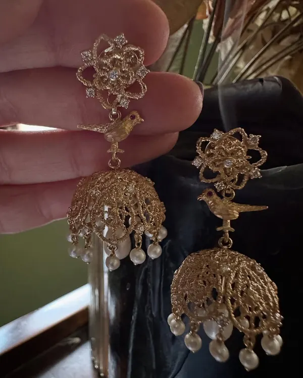 A close up of my handcrafted jhumka earrings. The design features a delicate bird motif and intricate filigree work in 18k gold, creating a classic dome shape adorned with pearls and diamonds.