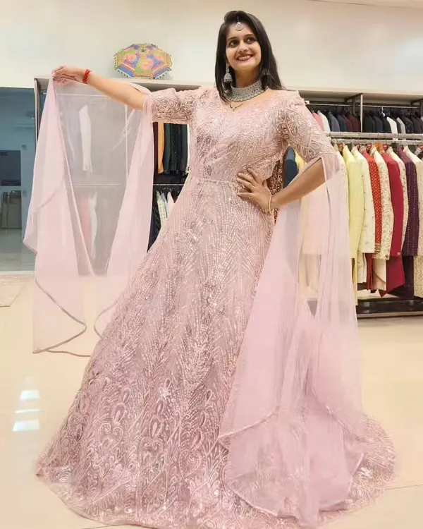 A full view of the stunning pink embroidered gown, showing its beautiful flare and the way the light catches the sequins. The attached cape-like sleeves add a touch of drama.