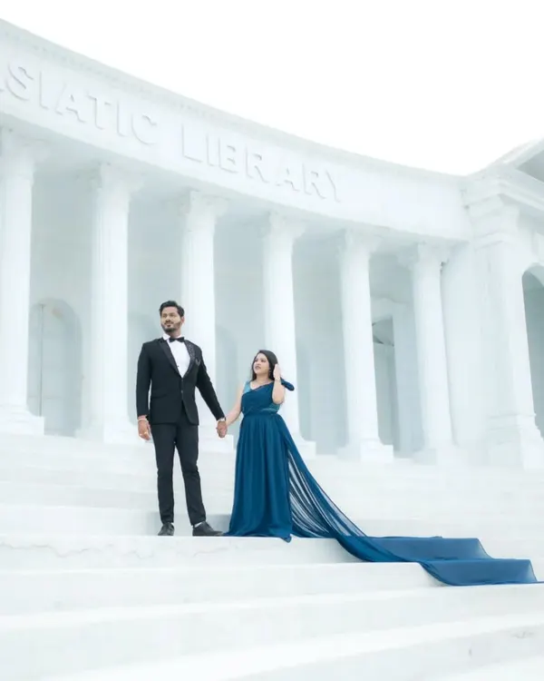 A majestic wide shot of a couple standing on the steps of an Asiatic Library replica set. The groom in a sharp tuxedo and the bride in a gown with a long train create a truly grand and aspirational pre-wedding portrait.