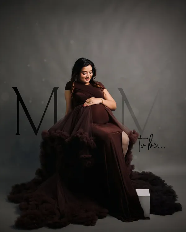 A classic and elegant studio portrait. The mother-to-be is seated in a dramatic, ruffled brown gown, looking down at her bump with a gentle smile, embodying the grace of this special time.