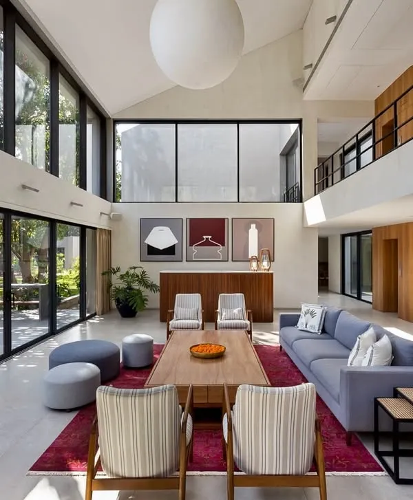 The living area of the Mango Orchard House is a study in volume and light. The double-height ceiling and expansive glass walls create a powerful connection to the outdoors, while the furniture arrangement fosters intimate conversation zones within the grand space.