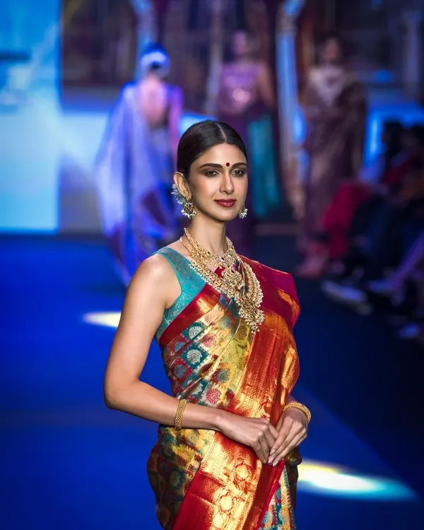 A classic runway shot from Bangalore Times Fashion Week, capturing the model's confident walk. The focus is on the traditional silk saree, showcasing its vibrant colors and elegant drape under the stage lights.