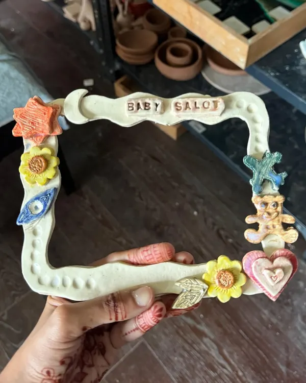 A close-up of a child's creation: a colorful, hand-built ceramic photo frame decorated with stars, flowers, and a teddy bear.