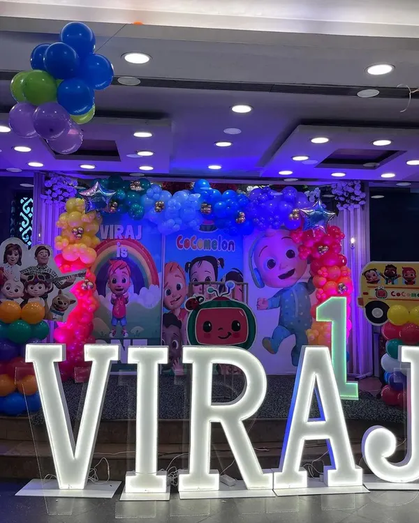 A vibrant Cocomelon stage setup for Viraj's first birthday. The decor features a large character backdrop, rainbow balloon columns, and light-up marquee letters for the name.