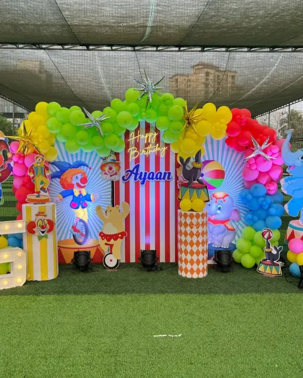 A spectacular outdoor circus carnival theme for Ayaan's 5th birthday. The setup includes a striped backdrop, colorful balloon arch, and cutouts of clowns, circus animals, and a large marquee number.