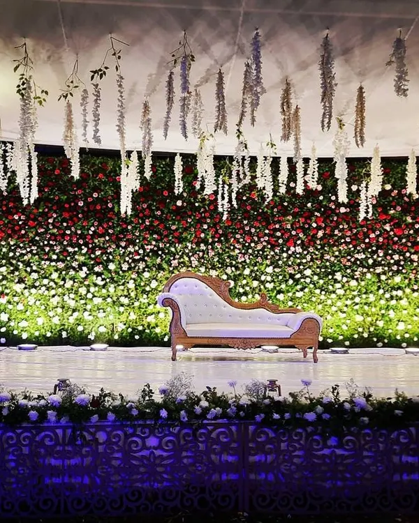 This outdoor reception stage features a massive wall of red and white flowers, creating a dramatic and romantic backdrop. I used hanging white floral strings from the ceiling and spotlights on the greenery pillars to add depth and texture to the design.