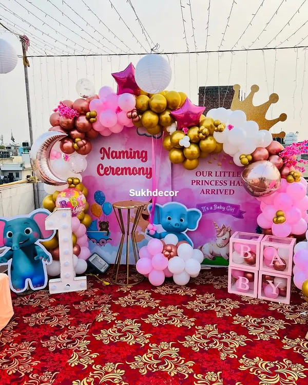 A royal welcome for a little princess. This naming ceremony on a rooftop features a large storybook-style backdrop, a pink, gold, and white balloon arch, and cute elephant cutouts.