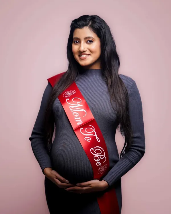 A classic studio maternity portrait. The simple pink background and the "Mom To Be" sash keep the focus on the expectant mother's happy expression.
