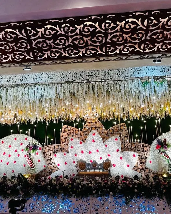 A grand reception stage featuring a lotus-inspired design made with white and red flowers. The cascading floral ceiling and intricate lighting create a truly breathtaking effect for a wedding reception.