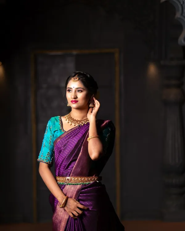 A dramatic portrait featuring a beautiful purple Kanchipuram silk saree. The lighting is designed to sculpt the subject and make the traditional jewelry and fabric shine.