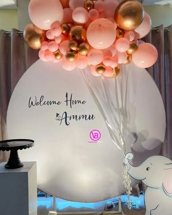 A sweet and charming setup to welcome a new baby home. This decoration includes a personalized "Welcome Home" backdrop, a pink and gold balloon arrangement, and a cute elephant cutout.