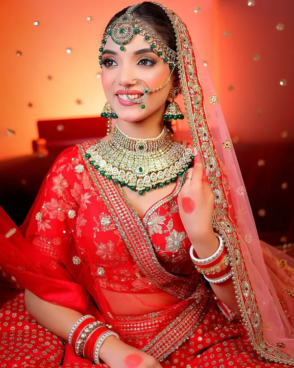 A smiling bridal portrait with a warm, celebratory background. The focus is on the intricate green and gold jewelry and the bride's happy expression.