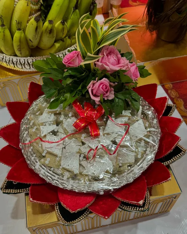 A classic sweet platter featuring Kaju Katli, presented on a silver tray with a red velvet lotus base. The arrangement is topped with fresh pink roses for an elegant touch.