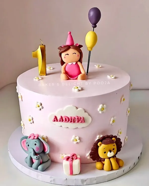 A pastel pink first birthday cake for little Aadhira. The design features cute fondant animals, a personalized name plaque, and a number '1' candle, making it a sweet and memorable centerpiece.