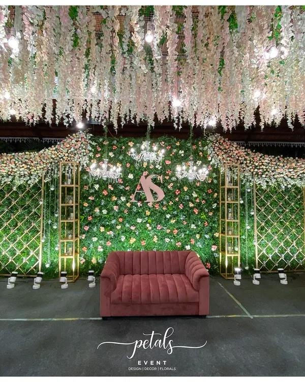 A luxurious reception stage featuring a dense green wall dotted with pastel flowers. A plush velvet sofa, custom couple's monogram, and a ceiling of hanging white florals create a look of pure opulence.