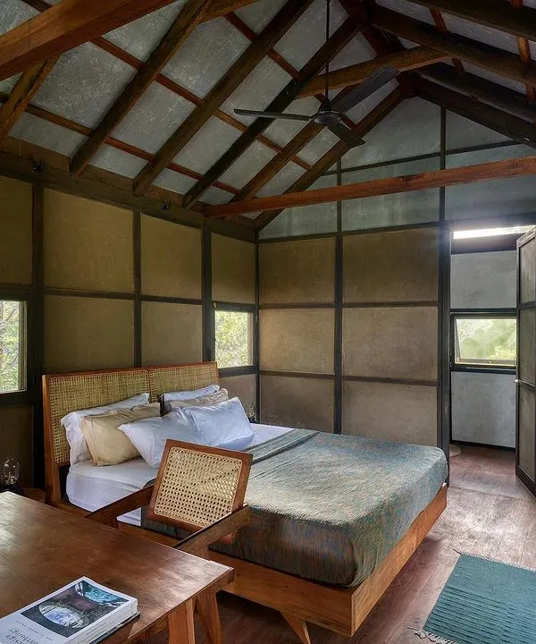 The bedroom of the prefab earthen prototype cottage. The design uses a simple palette of wattle-and-daub walls, a wooden frame, and an exposed timber roof to create a warm, serene, and rustic space.