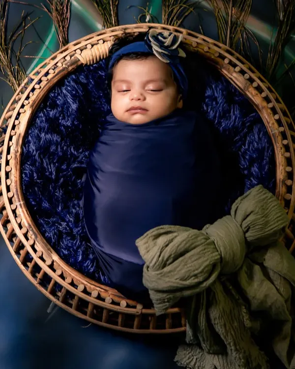 A peaceful newborn swaddled in royal blue, nestled in a rustic basket surrounded by peacock feathers. This is a classic example of a creative newborn setup.