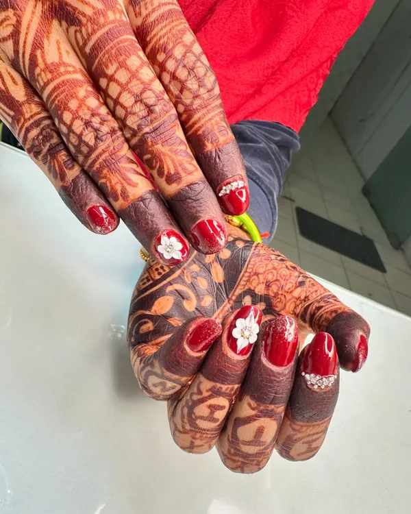 A stunning set of bridal nails designed to perfection. The deep red polish is accented with 3D white flowers and sparkling stones, creating a beautiful contrast with the intricate mehndi.