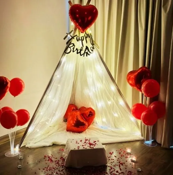 A romantic and intimate birthday surprise created right in the bedroom. This cozy setup features a beautiful teepee tent draped in sheer fabric and fairy lights, with red heart-shaped balloons and scattered rose petals to create a special, personal celebration.