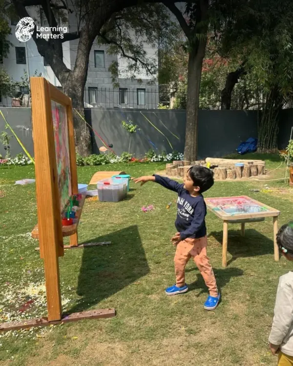 Our outdoor space becomes a large-scale art studio. Here, a child throws a paint-soaked ball at a clear easel, learning about trajectory, force, and color mixing in a dynamic, full-body engagement.