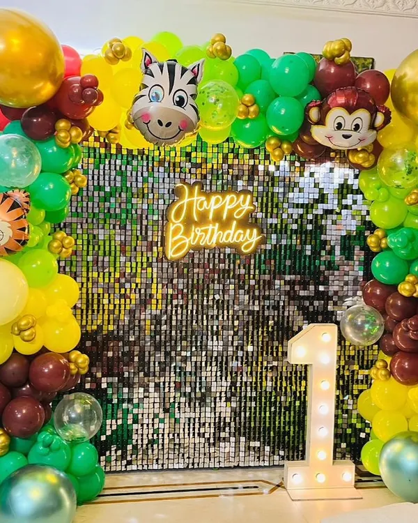 A jungle-themed first birthday party backdrop. This setup features a shimmering sequin wall, a 'Happy Birthday' neon sign, a light-up number '1', and a balloon arch with cute animal faces.