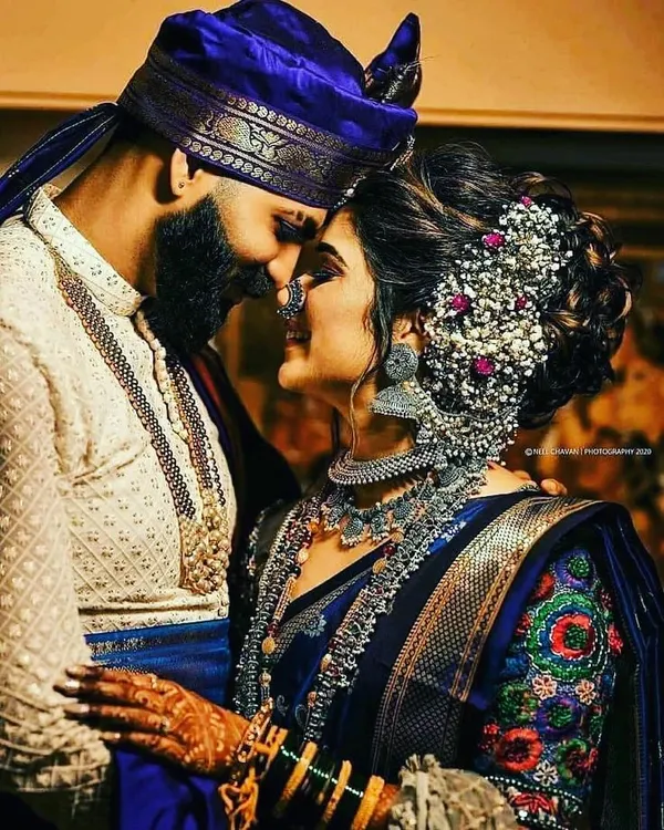 A beautiful moment between a bride and groom. The groom is wearing a traditional off-white sherwani with a royal blue stole and a matching blue safa, a classic Maharashtrian groom look.