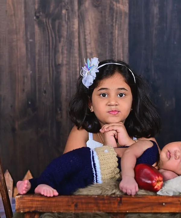 A big sister proudly watches over her sleeping newborn brother. This rustic studio setup with a wooden backdrop adds a warm, timeless feel to this sweet sibling portrait.