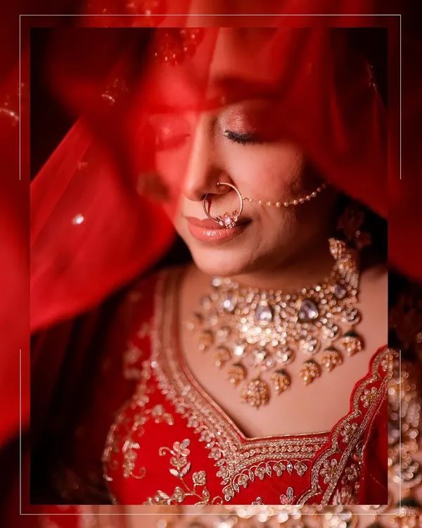 A serene and artistic bridal portrait. The bride's face is partially veiled by her red dupatta, drawing attention to her beautiful nose ring, intricate necklace, and peaceful expression.