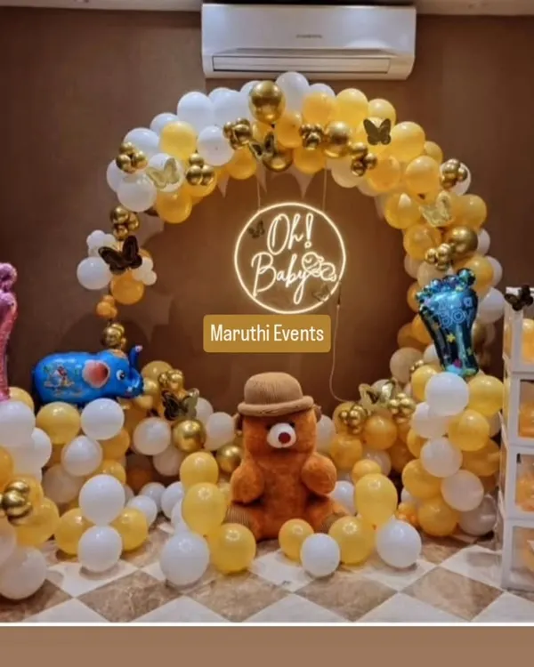 A charming 'Oh Baby' shower decoration with a yellow and white balloon arch, a neon sign, and cute teddy bear and elephant figures. The transparent 'BABY' blocks add a modern touch.