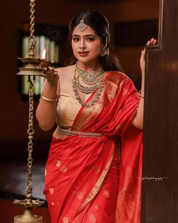 A classic portrait of a model in a red saree and gold blouse, holding a traditional lamp. The warm lighting and heritage setting create an atmosphere of timeless Tulunadu elegance.