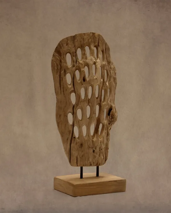 This handcrafted wood artifact features a unique perforated design, playing with light and shadow. Mounted on a simple teak base, it stands as a distinctive accent for any interior.