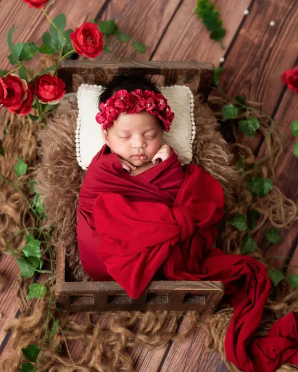 A timeless portrait of a newborn girl, peacefully sleeping in a miniature wooden bed. She is swaddled in a rich red wrap with a matching floral headband, surrounded by delicate red roses and ivy against a rustic wood backdrop.