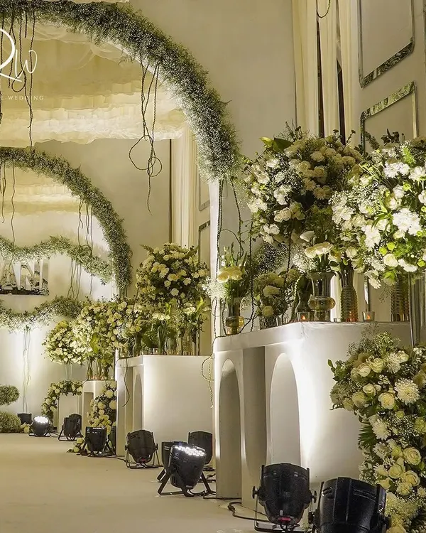 An enchanted forest theme brought to life. This setup features a walkway lined with white structures and massive arrangements of white flowers and greenery, with hanging arches of baby's breath.