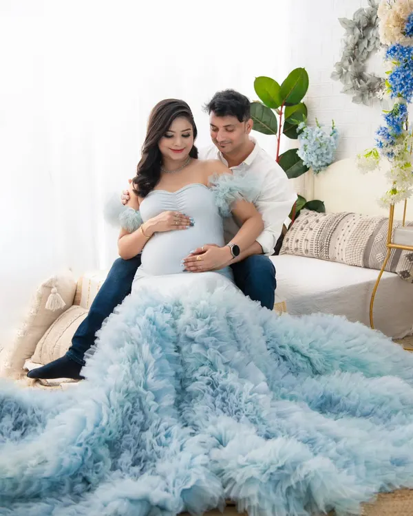 An intimate moment shared between the couple. The way they look at each other while cradling the bump speaks volumes about their shared love and excitement. The soft blue gown and light-filled setting create a serene atmosphere.