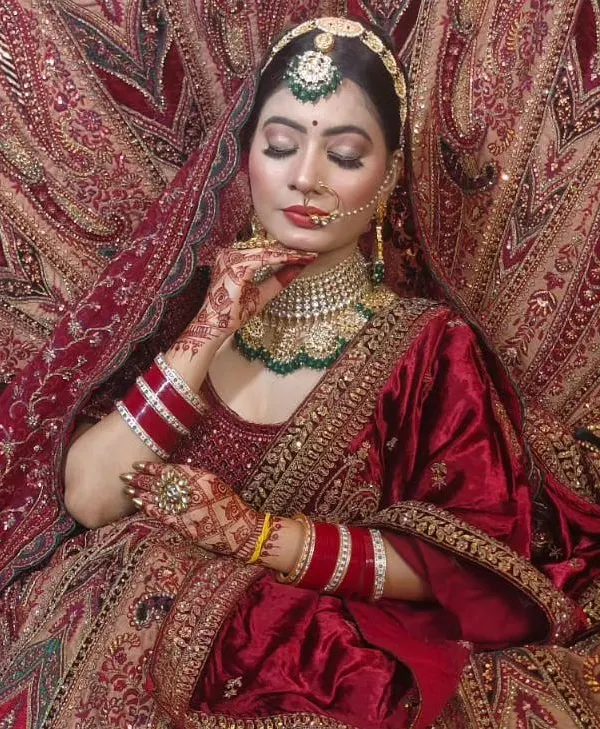 A regal bridal portrait in a deep red lehenga. The model is framed by the rich fabric, showcasing the intricate gold embroidery and a heavy choker set with green accents.