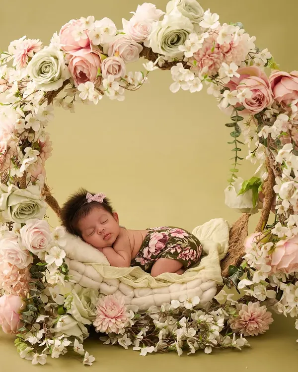 A newborn girl sleeps peacefully in a basket swing, completely encircled by a magnificent wreath of pink and white roses, creating a stunning floral masterpiece.